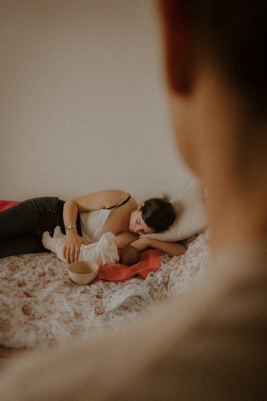 une jeune maman allaite son nouveau-ne allonges sur le lit sous le regard du papa pendant seance photo a leur domicile