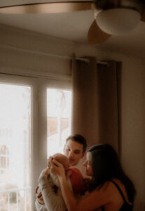 un jeune papa porte son nouveau-ne contre son epaule la maman le regarde en lui tenant la tete devant la fenetre de leur domicile